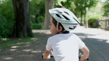 Parkta kaskla bisiklete binen bir çocuk, arkadan görülüyor. Çocuk yana bakıyor, güneşli bir günün tadını çıkarıyor ve yeşil ağaçlarla çevrili açık hava oyununun özgürlüğünü yaşıyor.