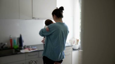 Anne mutfakta birden fazla iş yapıyor, yemek hazırlarken bebeğini kucağında tutuyor, camdan içeri ışık süzülüyor, ev ortamında modern ebeveynliğin zorluklarını ve güzelliklerini vurguluyor.