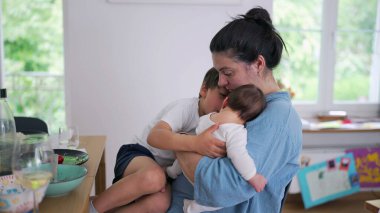 Anne hem bebeği hem de küçük bir çocuğu kucağında tutuyor, sıcak bir şekilde gülümsüyor. Kapalı bir ortamda ebeveynliğin neşeli ve besleyici tarafı.