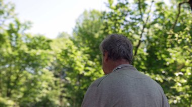 Yeşil ağaçlar ve doğa ile çevrili yaşlı bir adam parkta bir patikada yürüyor. Arkaya bakınca gri bir kazak giydiğini, açık havada huzurlu ve güneşli bir günün tadını çıkardığını görüyoruz.