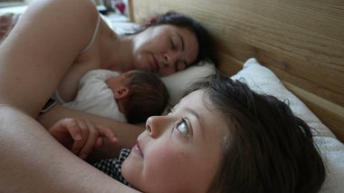 Anne ve küçük oğul yatakta, oğul düşünceli görünüyor anne ve yeni doğan uyurken, sabahın erken saatlerinde bağ kurma, aile birlikteliği, bakım, anne şefkati, samimi ve samimi bir an.