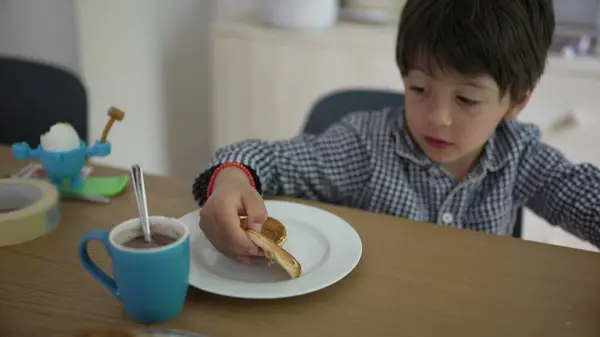 stock image Young boy sitting at a table, holding a pancake and looking at it with curiosity. The scene captures a moment of exploration and enjoyment during breakfast in a cozy home setting