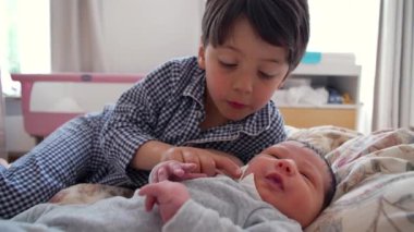Ağabeyi, yeni doğan kardeşiyle yatakta etkileşim halinde. Sıcak, aile odaklı bir evde kardeşler arasındaki özel bağ ve ilgiyi vurguluyor.