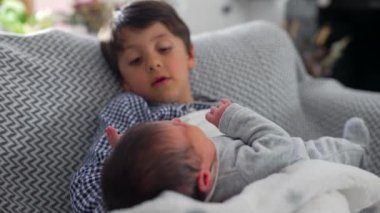 Büyük kardeş, sıcak bir aile ortamında derin bir kardeşlik bağı kurmaya başladıklarında, onları nazikçe tutarken, bir anlık merak ve yakınlık göstererek, yeni doğan kardeşe dikkatle bakıyor.