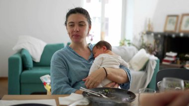 Anne yemek çiğnerken kucağında bebeği, sıcak bir oturma odasında oturuyor. Ebeveynliğin çok yönlü bir özelliği, huzurlu bir ev ortamı ve sakin, besleyici bir atmosferi var.