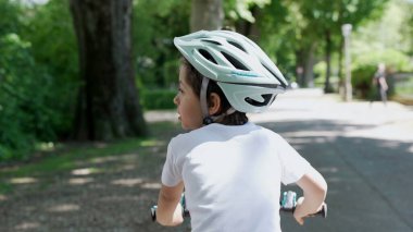 Parkta kaskla bisiklete binen bir çocuk, arkadan görülüyor. Çocuk yana bakıyor, güneşli bir günün tadını çıkarıyor ve yeşil ağaçlarla çevrili açık hava oyununun özgürlüğünü yaşıyor.