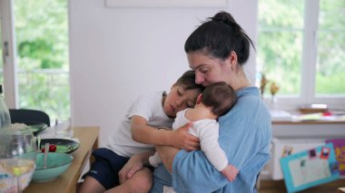 Anne hem bebeği hem de küçük bir çocuğu kucağında tutuyor, sıcak bir şekilde gülümsüyor. Kapalı bir ortamda ebeveynliğin neşeli ve besleyici tarafı.