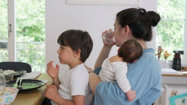 Anne bir bebeği tutarken su içiyor ve başka bir çocuğun yemek yemesini seyrediyor. Bu sahne anne olmanın çoklu görev ve şefkatli doğasını bir ev ortamında yakalar.