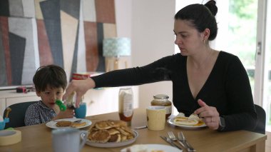 Anne ve oğlu ahşap bir masada kahvaltı yapıyor, anne çocuğun kreplerinin üzerine şurup döküyor, sıcak ve aileye yönelik bir atmosfer yaratıyor.