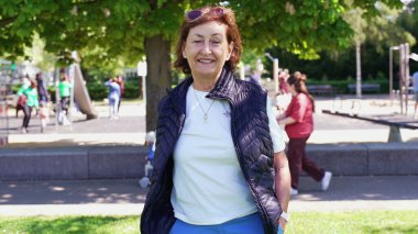 Yaşlı bir kadının siyah yelek ve beyaz gömlek giyerek parkta duruşu. Arka planda bir oyun alanı ve günün tadını çıkaran insanlar var.