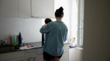 Anne mutfakta birden fazla iş yapıyor, yemek hazırlarken bebeğini kucağında tutuyor, camdan içeri ışık süzülüyor, ev ortamında modern ebeveynliğin zorluklarını ve güzelliklerini vurguluyor.