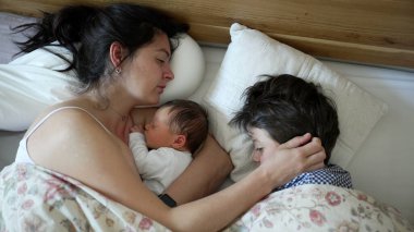 Anne yeni doğmuş bebeği ve küçük çocuğuyla rahat bir yatakta huzur içinde uyuyor, aile bağı ve şefkat, sevgi dolu anne bebeğe sarılıyor, büyük kardeş sarılıyor, sabah rutini, huzur içinde dinleniyor.