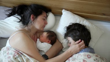 Anne yeni doğmuş bebeği ve küçük çocuğuyla rahat bir yatakta huzur içinde uyuyor, aile bağı ve şefkat, sevgi dolu anne bebeğe sarılıyor, büyük kardeş sarılıyor, sabah rutini, huzur içinde dinleniyor.