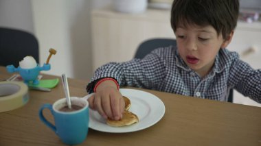 Genç bir çocuk masada oturuyor, elinde bir krep tutuyor ve ona merakla bakıyor. Sahne, kahvaltı sırasında sıcacık bir ev ortamında keşif ve keyif anı yakalıyor.