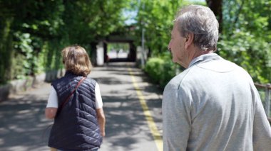 Ağaçlık bir yolda yürüyen yaşlı bir çiftin, güneşli bir günün tadını çıkarışının arka görüntüsü. Yeşillik ve benekli güneş ışığıyla çevrili, emekli yaşlılıkta düşünceli yürüyüş.