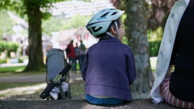 Motosiklet kaskı takan bir çocuk parkta annesinin yanında oturuyor, scooter bankta dinleniyor, aile açık havada güneşli bir günün tadını çıkarıyor, güvenlik ve rahatlama için sıradan bir sahne.