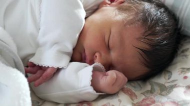 Koyu renk saçlı yeni doğmuş bebek çiçek yatağında mışıl mışıl uyuyor, kolları içeri sokulmuş, sıcaklıkla dolu sıcak ve güvenli bir ortamda ilk yaşamın şefkatini ve huzurunu sergiliyor.