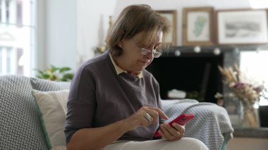 Gözlüklü yaşlı bir kadın koltukta otururken kırmızı akıllı telefon kullanıyor, teknolojiyle meşgul, rahat ev dekorasyonu yapıyor, modern cihazlar ve iletişim araçlarıyla üst düzey iletişimi vurguluyor.