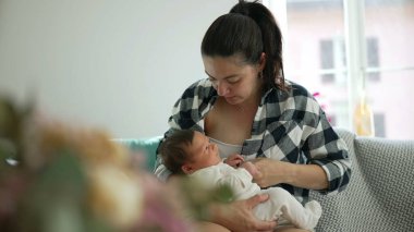 Anne bebeğini emzirmek üzere, kendini yeni doğan bebeğine süt vermeye hazırlıyor, rahat bir ev ortamında, rahat bir kanepede oturuyor, güçlü bir sevgi, ilgi ve annelik duygusu var.