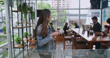 Kot ceketli gülümseyen kadın kahve dükkanındaki müşteriden kahve fincanı alıyor. Arka planda çalışan ve sosyalleşen insanların olduğu parlak, hoş bir ortam.