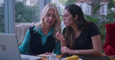 İki kadın bir kafede dizüstü bilgisayarda bir şey tartışıyor. Bir kadın sarı saçlı ve mavi bluzlu, diğeri uzun siyah saçlı, ikisi de işbirlikçi bir konuşma içinde.
