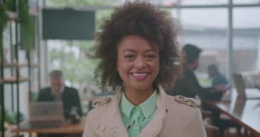 Modern bir kahve dükkanında açık renk ceketli, kıvırcık saçlı, gülümseyen bir kadın. Arka planda çalışan ve sosyalleşen insanların olduğu parlak atmosfer canlı ve davetkar bir ortamı vurguluyor.