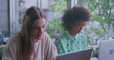 Kahve masasında dizüstü bilgisayarlarla çalışan iki kadın, ikisi de derin düşüncelere dalmış. Görüntü odaklanmayı, üretkenliği ve davetkâr bir ortamda profesyonel ama gelişigüzel bir çalışma alanını vurgular