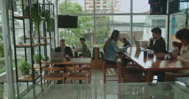 Laptoplarla çalışan ve sohbet eden insanlarla dolu bir kafe. Kahve ekipmanları sıcak bir ortam ekler. Geniş pencereler doğal ışık girmesini sağlıyor, parlak ve verimli bir atmosfer yaratıyor.
