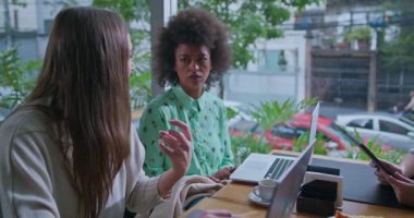 Üç kadın dizüstü bilgisayarlarla ve bir tabletle pencerenin yanındaki kafede çalışıyor. Sahne, rahat bir atmosferi olan, işbirlikçi, kentsel çalışma alanındaki verimlilik ve teknoloji kullanımını vurguluyor