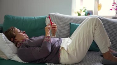 Akıllı telefon kullanırken kanepede dinlenen yaşlı bir kadın, sessiz boş zamanların tadını çıkarıyor ve teknoloji ile bağlı kalıyor, modern rahatlığı rahat bir ev ortamında harmanlıyor.