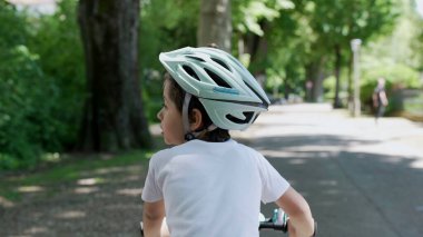 Genç çocuk parkta kaskla bisiklet sürüyor, arkasını görüyor güneşli bir günün keyfini çıkarıyor, açık hava aktivitesi, ağaçlarla çevrili, doğayı keşfediyor, çocuk güvenliği, günlük kıyafet, çocukluk macerası.