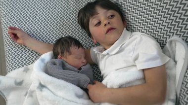 Genç çocuk, her ikisi de yumuşak bir battaniyeye sarılı uyuyan yeni doğan kardeşini sevgiyle izliyor ve kardeşler arasındaki hassas ve koruyucu anı yakalıyor.