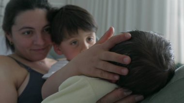 Anne ve büyük kardeş yeni doğan bebeğin kafasına sevgi ve özenle dokunuyor. Hastane yatağında hassas anlar aile içindeki nazik etkileşimi yakalar ve yeni bir bebeğe kucak açmanın sevincini vurgular.