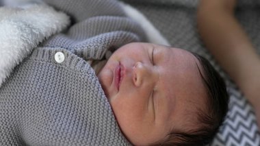 Yeni doğmuş bir bebeğin, örgü örülmüş gri bir kıyafetle huzurlu bir şekilde uyuduğu, narin yüz hatlarını ve huzurlu ifadesini vurguladığı, rahat bir battaniyeye sarılı olduğu yakın plan.