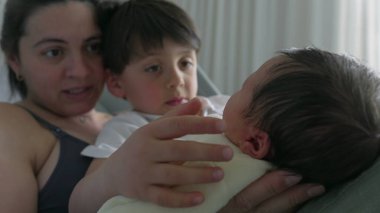 Anne ve büyük kardeş yeni doğan bebeğin kafasına sevgi ve özenle dokunuyor. Hastane yatağında hassas anlar aile içindeki nazik etkileşimi yakalar ve yeni bir bebeğe kucak açmanın sevincini vurgular.