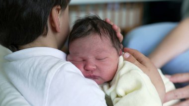 Ağabeyi kucağında uyuyan yeni doğmuş bir bebeği kucaklıyor, kardeş sevgisini, koruyucu bağını ve hastane ortamında aile bağının hassas anlarını vurguluyor.