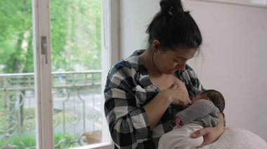 Anne, yeni doğmuş bebeğini sıcak, güneşli bir odada sevgiyle emziriyor ve hassas bir bağ ve bakım anı yaşıyor. Bebek annesinin kollarında rahatça besleniyor.