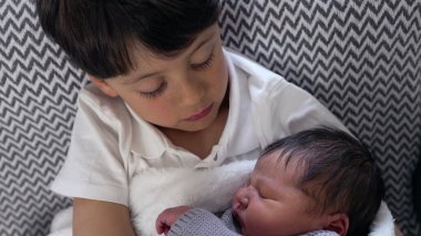 Genç çocuk, sıcak bir battaniyeye sarılmış yeni doğan kardeşine sevgiyle bakar. Kardeşçe bir yakınlık ve aile bağının hassas bir anını yakalar.