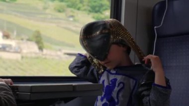 Şövalye kostümü giymiş genç bir çocuk trende giderken kaskını takıyor, ortaçağ fantezisinin eğlenceli unsurlarını modern bir yolculukla harmanlıyor, odaklanmış bir hayal gücü anı.