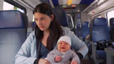 Anne, beyaz şapkalı bebeği trende tutarken kameralar önünde biriyle konuşuyor. Annelik ve toplu taşıma sırasında günlük yaşamın dengesini vurguluyor.