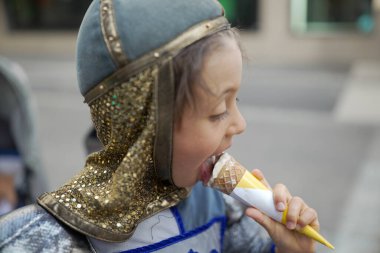 Ortaçağ şövalyesi kostümlü bir çocuk festivalde vanilyalı dondurma yiyor. Yan profil, çocuğun zevkini ve tarihi kıyafetlerin modern bir ziyafetle harmanlanmasını gösteriyor.