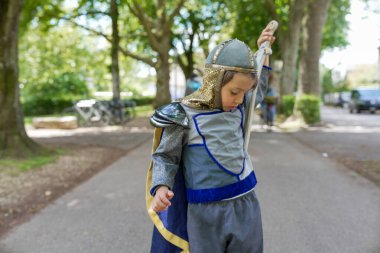 Şövalye kostümü giymiş küçük bir çocuk elinde kılıçla dışarıda yürüyor ve eğlenceli bir maceranın tadını çıkarıyor.