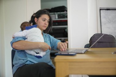 Anne kucağında bebek, çoklu görev, mavi yakalı beyaz tulumlu bebek, kapalı mekan sahnesi, denge işi ve annelik, modern aile hayatı, odaklı ifade