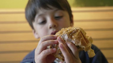 Çocuk, heyecanına ve yemeğin dokusuna odaklanmış lezzetli bir çizburgerin tadını çıkarıyor. Çocukluk sevincini ve lezzetli bir yemek yemenin basit zevklerini yakalamak için mükemmel bir an.