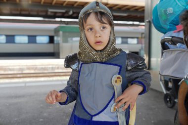 Bir şövalye gibi giyinmiş bir çocuk tren istasyonunda bekliyor, elinde bir kılıç ve garip bir ifadeyle bir kask taşıyor.