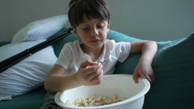 Küçük çocuk kanepede patlamış mısır yiyor, kendini bir şeyler izlemeye kaptırmış, çocukluğunun samimi bir anını yakalıyor.