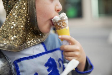 Ortaçağ şövalyesi kostümü giymiş bir çocuk açık hava festivalinde dondurma külahının tadını çıkarıyor. Eğlenceli sahne, tarihi canlandırma ile günlük eğlenceyi birleştirir. Çocuğun zevkini gözler önüne serer.