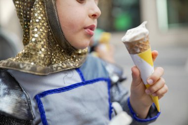 Ortaçağ şövalyesi kostümlü bir çocuk festivalde vanilyalı dondurma yiyor. Yan profil, çocuğun zevkini ve tarihi kıyafetlerin modern bir ziyafetle harmanlanmasını gösteriyor.