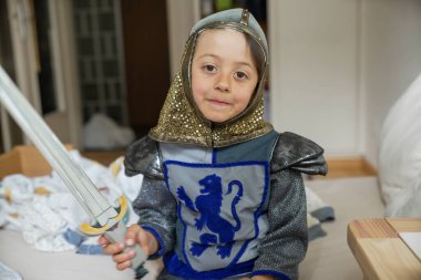 Küçük çocuk oyuncak kılıçlı bir şövalye gibi giyinmiş, yatakta oturuyor, yaratıcı bir oyun, ortaçağ kostümü giyiyor, yatak odası sahnesi, çocukluk, yaratıcılık, neşeli bir ifade, şövalye zırhı giyiyor.