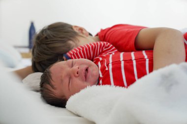 Yeni doğan bebek ağlarken, büyük kardeş yatakta yatarken, kırmızı kıyafetler içinde, duygusal bir anı yakalıyor, kardeş ilişkilerinin zorluklarını ve bağlarını vurguluyor.